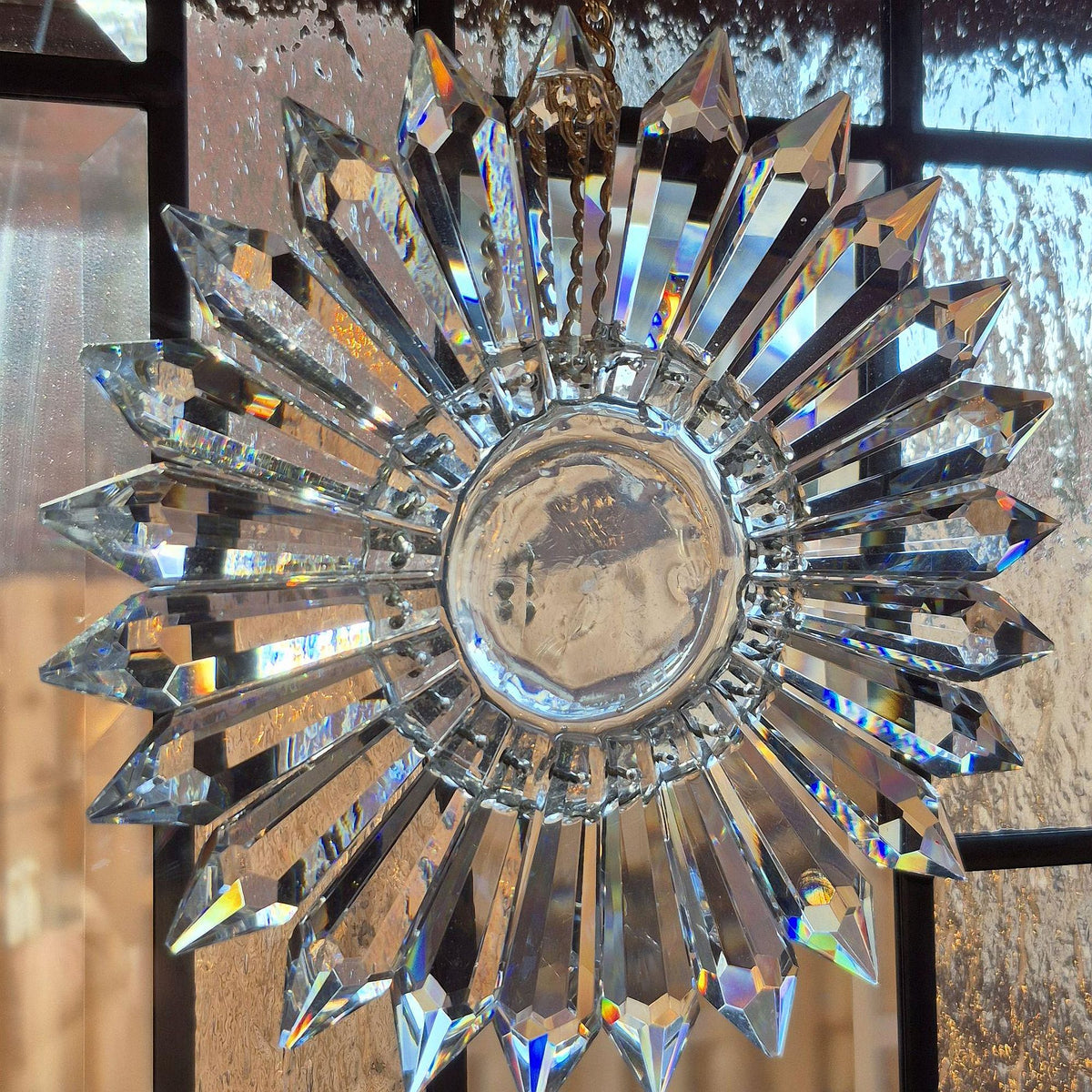 A clear crystal prism suncatcher in a radial pattern hanging from a chain, featuring multiple faceted star-shaped prisms radiating from a round central base over Art Deco divided light window